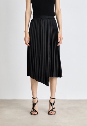 Femme portant un haut noir sans manches, une jupe midi plissée asymétrique noire et des sandales noires à talons hauts à brides, debout sur un sol gris.