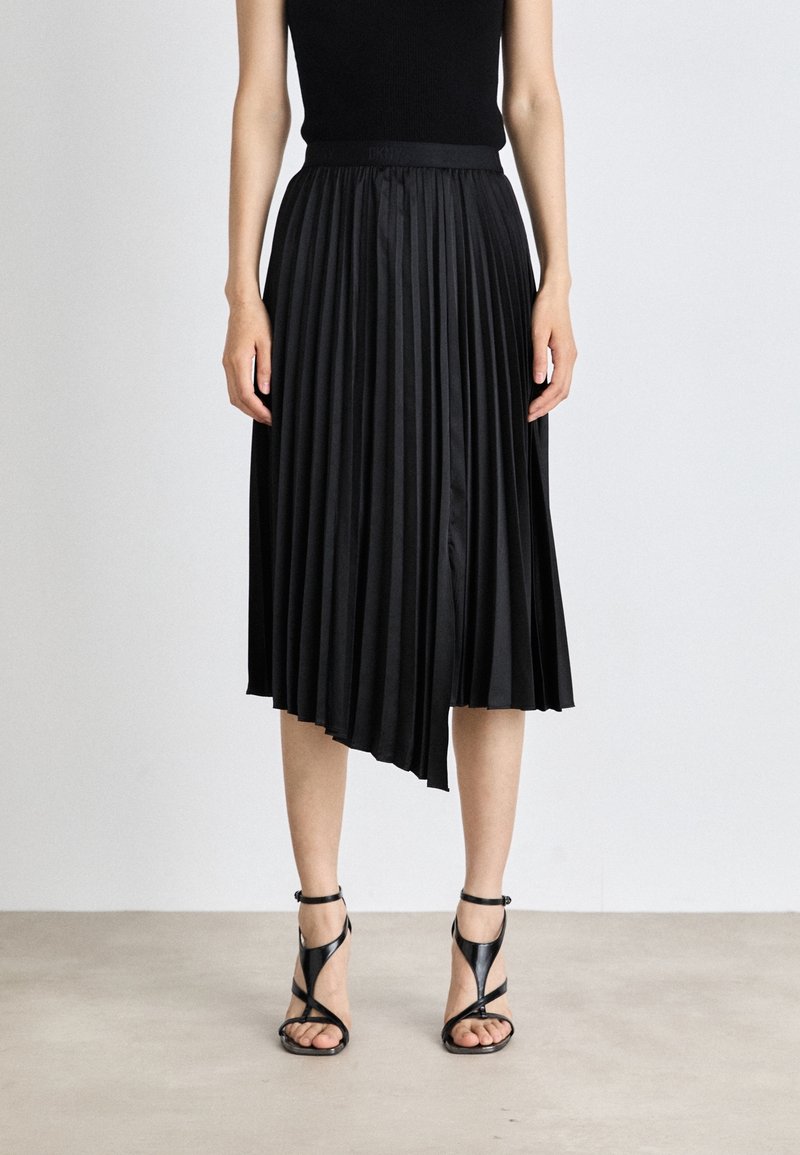 Femme portant un haut noir sans manches, une jupe midi plissée asymétrique noire et des sandales noires à talons hauts à brides, debout sur un sol gris.