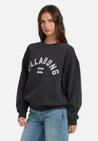 Czarny, oversize'owy sweatshirt z logo "Billabong" w białym kolorze, z prążkowanymi mankietami i okrągłym dekoltem, w zestawieniu z jasnoniebieskimi dżinsami.