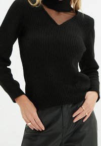 Schwarzer Strickpullover mit V-Ausschnitt und hohem Kragen. Ripptextur, taillierte Silhouette und lange Ärmel. Kombiniert mit schwarzen Lederhosen.