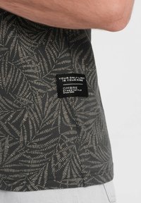 Košile s krátkým rukávem z tmavě šedé látky s listovým béžovým vzorem. Obsahuje štítek s textem, který uvádí značku a slogan.