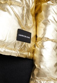 Gouden donsjack met een glanzende afwerking, voorzien van een zwart label met de tekst "CALVIN KLEIN JEANS" en zichtbare stikdetails.