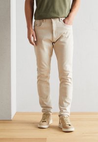 Vaqueros de mezclilla beige con corte ajustado, diseño de cinco bolsillos y cierre de botón, combinados con zapatillas beige y una camiseta verde de cuello redondo.