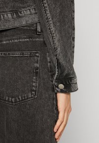 Černá džínová bunda s texturovaným povrchem, ozdobená patentkami na manžetách a kontrastním prošíváním. Viditelná boční kapsa, úzký střih.