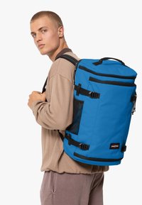 Blå ryggsäck med svarta detaljer, tillverkad av slitstarkt tyg. Har en nätficka, justerbara remmar och ett handtag på toppen för enkel bärning.