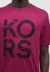 Άτομο που φοράει ματζέντα t-shirt με μεγάλα μαύρα γράμματα "KORS" τυπωμένα στο μπροστινό μέρος, τα οποία δείχνουν ένα διακριτικό μοτίβο φύλλου μέσα σε κάθε γράμμα.