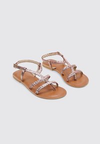 Sandalias planas en oro rosa con correas trenzadas y suela marrón. Diseño de punta abierta con una correa en el tobillo ajustable para un mejor ajuste. Superficie de textura suave.