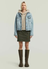 Giacca di jeans, felpa beige con cappuccio, gonna verde e stivali alti marroni. La giacca ha due tasche sul petto, la felpa presenta una tasca a marsupio, tessuto strutturato.