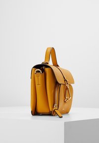 Żółty skórzany plecak o teksturowanym wykończeniu, z klapką zamykaną na zatrzask, boczną kieszenią na suwak oraz złotymi akcentami metalowymi. Kompaktowy i uporządkowany kształt.