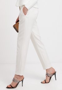 Vrouw in witte op maat gemaakte broek, met een bruine clutch, en zwarte sandalen met hoge hakken voorzien van een slangenhuidpatroon.