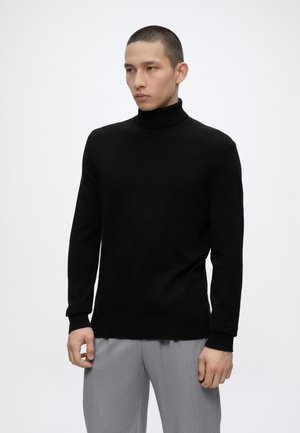 Hombre que lleva un suéter de cuello alto negro y pantalones gris claro, de pie frente a un fondo blanco liso, mirando ligeramente hacia la izquierda.