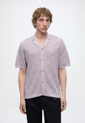 Jeune homme aux cheveux bouclés châtain clair portant une chemise en tricot lavande à manches courtes avec un motif à carreaux texturé.