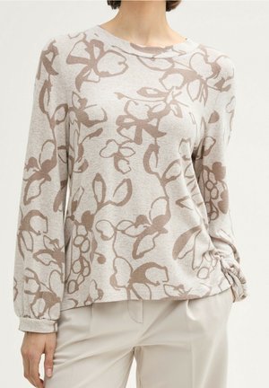 Pull beige à manches longues avec motif floral abstrait marron clair, porté avec un pantalon beige clair taille haute, main reposant près de la poche.