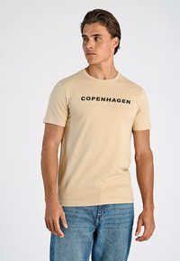 Beige t-shirt med kort ärm och rund halsringning, med en djärv svart "COPENHAGEN"-tryck över bröstet. Slät, åtsittande tyg.