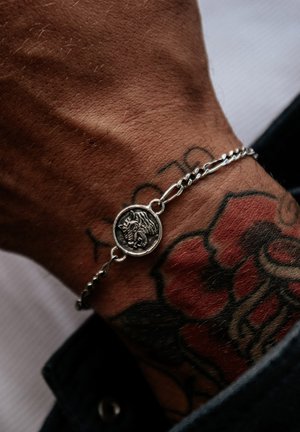 Silver chain bracelet featuring a round pendant with a lion design, worn on a hand with a visible tattoo. Textured hand and contrasting skin tone.