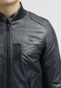 Mörkblå läderjacka med dragkedja framtill, ribbad krage och en liten svart logotyp. Har en texturerad yta och sömmar vid axlarna.