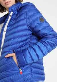 Blå polstret jakke med hætte, der har syede paneler, en lynlåslomme og et logo-patch på armen. Glat, skinnende stofstruktur.