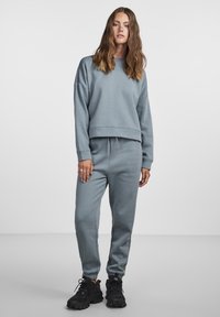 Lys grå hettegenser og joggebukse-sett, laget av mykt stoff. Har en kortet design, elastisk midje og oppbrettede ankler. Kombinert med svarte joggesko.