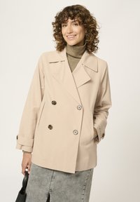 Chaqueta beige de doble botonadura con grandes botones, solapas anchas y corte relajado. La tela parece suave, combinada con un ligero cuello de tortuga.