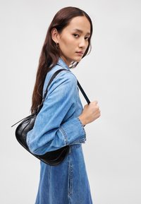 Μαύρη δερμάτινη τσάντα ώμου με καμπύλη μορφή, φορεμένη πάνω από ένα τζιν φόρεμα. Διαθέτει λεπτό, ρυθμιζόμενο λουρί και λεία υφή.