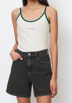 Kobieta ubrana w biały top na ramiączkach z zielonym obszyciem i czarne, jeansowe szorty z wysokim stanem, z jedną ręką w kieszeni na białym tle.