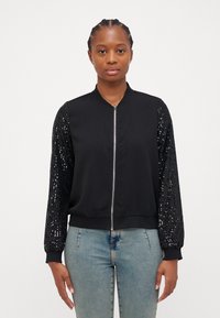 Veste bomber noire avec un corps lisse et des manches longues couvertes de sequins ; elle possède une fermeture éclair à l'avant et des poignets et un ourlet côtelés.