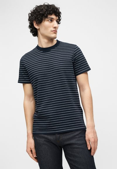 Νεαρός με κατσαρά μαλλιά που φοράει μπλε ναυτικό μπλουζάκι με λεπτές λευκές οριζόντιες ρίγες και σκούρο τζιν, κοιτάζοντας προς τα πλάγια.