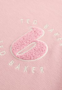 Vaaleanpunainen tekstuuri kangas, jossa on suuri, pörröinen vaaleanpunainen brodeerattu kirjain "B" valkoisella reunuksella, ympärillä valkoiset tekstit "TED BAKER."