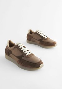 Brune ruskind- og lædersneakers med cremefarvede snørebånd, rund tå og teksturerede detaljer. Lys beige gummisål giver støtte.