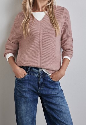Femme portant un pull côtelé rose poussiéreux par-dessus une chemise blanche, associé à un jean en denim bleu, les mains dans les poches, debout contre un mur uni.