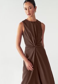 Vestido marrón sin mangas con un detalle anudado en la cintura, que presenta una textura suave y una silueta estructurada.