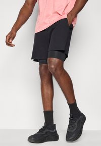 Μαύρα αθλητικά σορτς με λείο ύφασμα, με στρωματοποιημένο σχέδιο και πλαϊνές τσέπες, συνδυασμένα με μαύρες κάλτσες και αθλητικά παπούτσια, και ροζ μπλούζα.