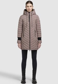 Kvinde iført en mauve, kviltet vinterfrakke med hætte, sorte tights og sorte ankelstøvler, stående foran en ensfarvet lys baggrund.