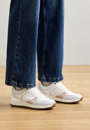 Sneakers bianchi con accenti beige, materiali lisci e testurizzati, punta rotonda e design con lacci, abbinati a jeans a gamba larga blu scuro.