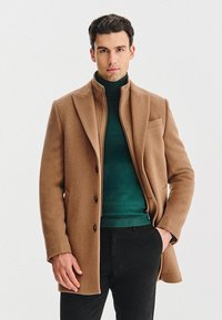 Brązowy wełniany płaszcz z klapami, wyposażony w kieszeń na piersi i trzy guziki. Pod spodem ciemnozielony sweter golf i czarne spodnie.