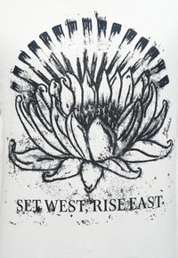 Σχέδιο με μαύρο μελάνι ενός ανθισμένου λουλουδιού με έντονα πέταλα πάνω από το κείμενο «ΑΝΑΤΟΛΗ ΑΝΑΤΟΛΙΚΑ, ΔΥΣΗ ΔΥΤΙΚΑ» σε λευκό ύφασμα.