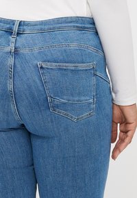 Jeans en denim bleu avec une taille mi-haute, présentant un design de poche arrière et des détails de couture subtils. La texture est lisse avec un léger élasticité.