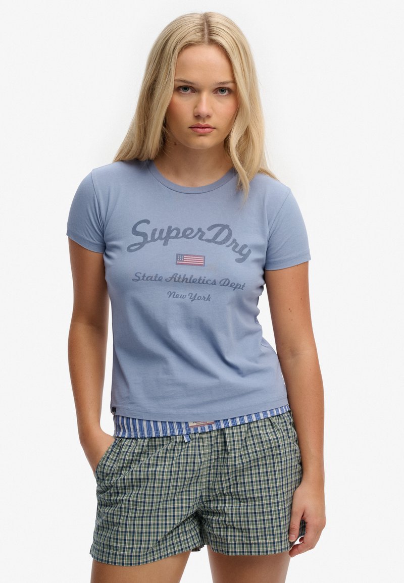 Mujer rubia con camiseta azul claro de "SuperDry" y pantalones cortos de cuadros verdes, de pie con una mano en el bolsillo contra un fondo blanco.