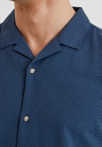 Navy blue short-sleeve shirt with a textured, waffle-like pattern, featuring a camp collar and a single metallic button detail.