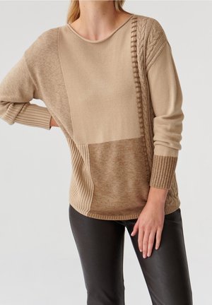 Femme portant un pull en maille patchwork beige avec un détail torsadé et un pantalon en cuir noir, debout sur un fond clair.