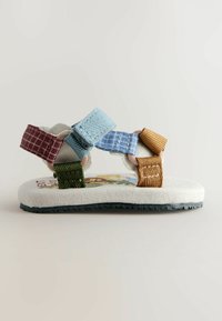 Sandale enfant avec des brides en tissu réglables multicolores en bleu, orange, vert et marron, et une semelle blanche légèrement imprimée.