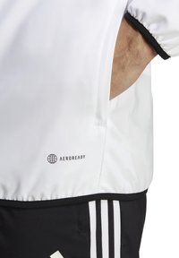 Chaqueta deportiva blanca con acentos negros, que presenta un dobladillo redondeado, puños elásticos y un logo "AEROREADY" cerca de la costura.