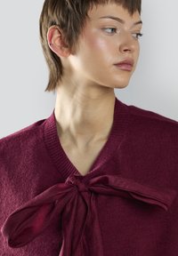 Maglione a collo a V in magenta con un grande dettaglio a fiocco in tessuto al collo. Texture morbida con rifiniture a coste al collo e alle maniche.