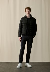 Black padded jacket with a high collar, worn over a black shirt and dark trousers. White sneakers complete the look. Smooth fabric texture.