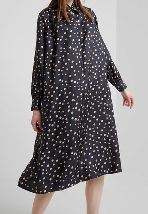 Nainen, joka käyttää löysää, mustaa pitkähihaista mekkoa, jossa on beigenvärisiä pilkkuja, seisoo yksinkertaisen vaaleanharmaan taustan edessä.