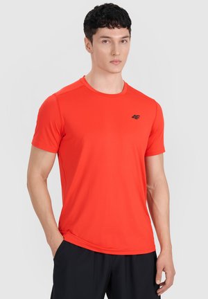Joven con cabello corto y oscuro que lleva una camiseta de manga corta de color rojo brillante con un pequeño logo negro en el pecho y pantalones negros.