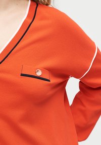 Γυναίκα που φοράει πορτοκαλί τοπ με λαιμόκοψη σε σχήμα V, με λευκή και μαύρη επένδυση, μικρή τσέπη με κουμπί στο στήθος και μακρυά μανίκια.