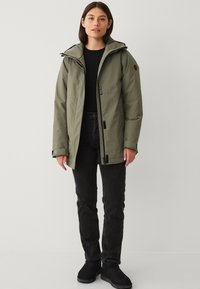 Donna con una giacca a cappuccio verde oliva, maglietta nera a maniche lunghe, jeans neri e stivali neri, in piedi davanti a uno sfondo neutro.