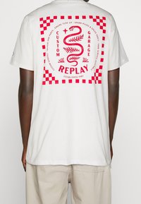 Bílý bavlněný tričko s velkým červeným grafikou s hadem a kostkovaným okrajem, s nápisem "REPLAY CUSTOM GARAGE ZALOŽENO 1981."
