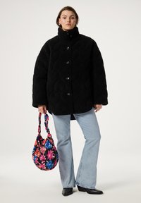 Zwarte, gestructureerde, oversized jas met een hoge kraag en knopen; gecombineerd met lichtblauwe flared jeans en een handtas met bloemmotief.